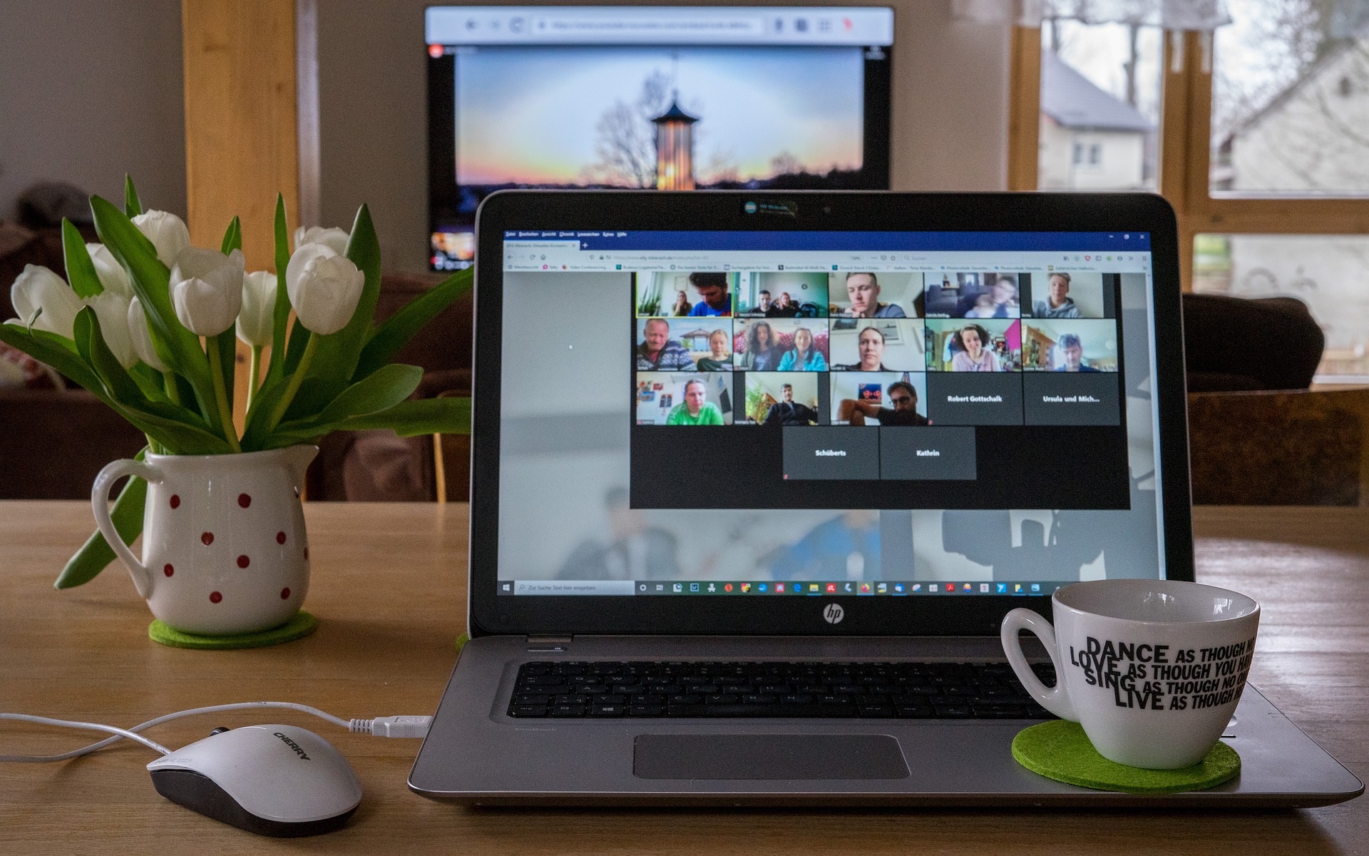 Ein Bild von einem Laptop, auf dem mehrere Personen in einem Online-Meeting sind. Es isnd eineige Personen mit Bild und in einigen Feldern steht ein Name. Rechts neben dem Laptop steht eine weiße Kaffeetasse, links daneben eine Vase mit weißen Tulpen. Links vor dem Laptop ist eine weißen Computermaus.